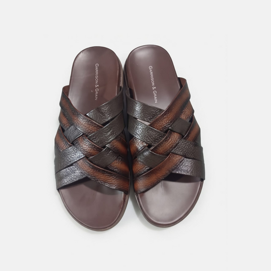 A pair of men's slide slippers with a braided woven leather upper in dark brown and black, set against a light gray background.